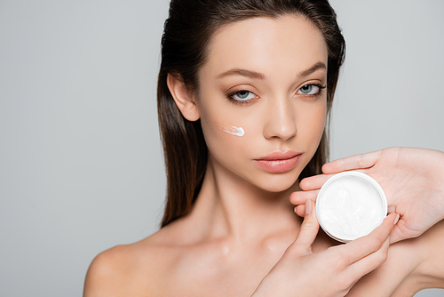 young woman with cosmetic cream on cheeks holding container isolated on grey