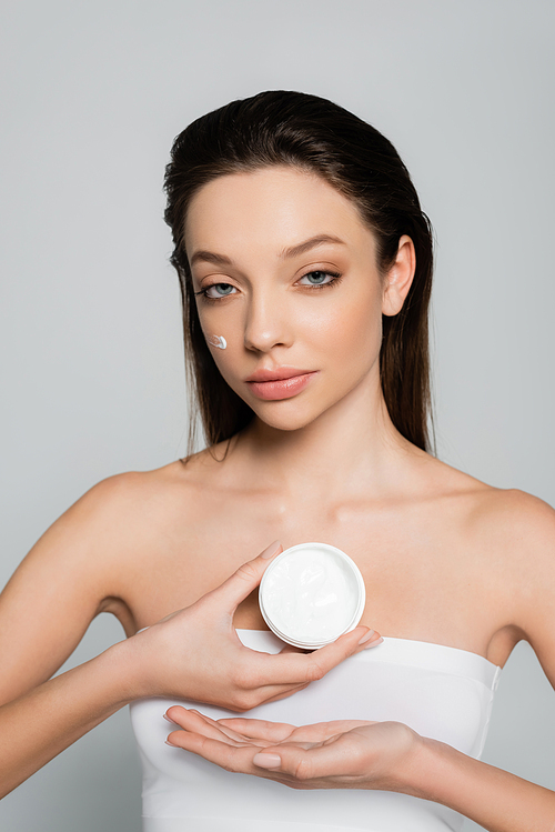 brunette young woman with bare shoulders and cream on cheeks holding container isolated on grey