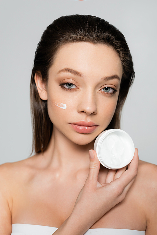 young woman with cream on cheeks holding container isolated on grey