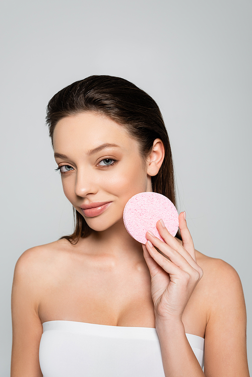 smiling young woman with bare shoulders holding exfoliating sponge isolated on grey