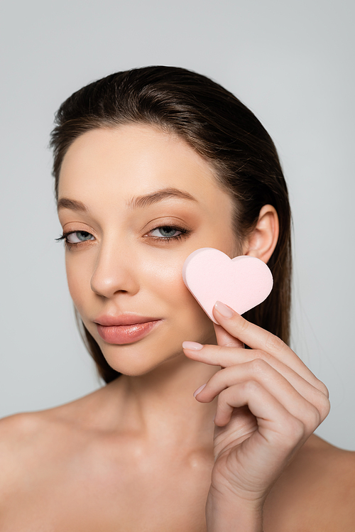 young woman holding heart-shaped sponge near face isolated on grey