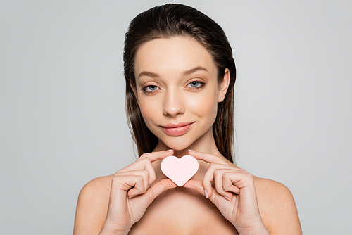 smiling young woman holding heart-shaped sponge near face isolated on grey