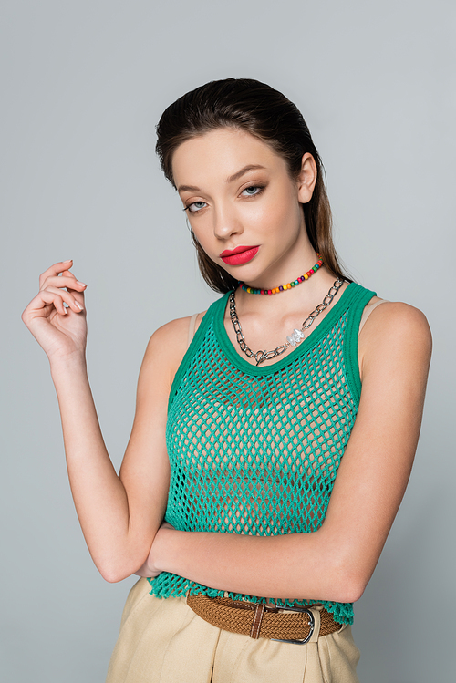 young and stylish woman with red lips and necklaces posing isolated on grey
