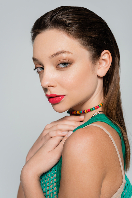 young woman with red lips touching colorful necklace isolated on grey