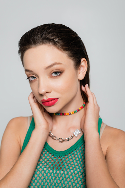 stylish model in green tank top and colorful necklace looking at camera isolated on grey