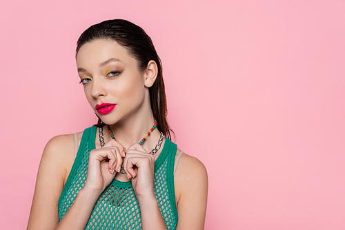 young brunette woman with bright makeup pulling necklaces while looking at camera isolated on pink