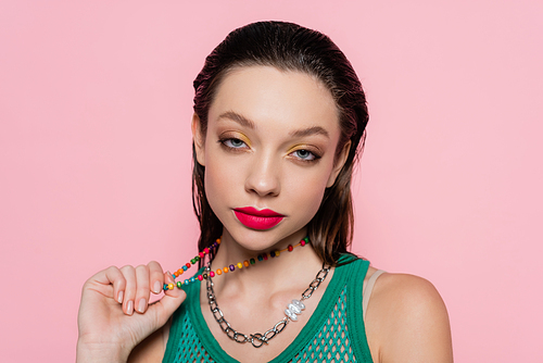 young brunette woman with bright makeup pulling beads necklace while looking at camera isolated on pink