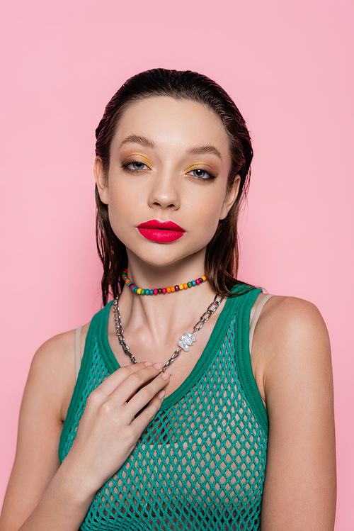 young brunette woman with bright makeup touching chain necklace while looking at camera isolated on pink
