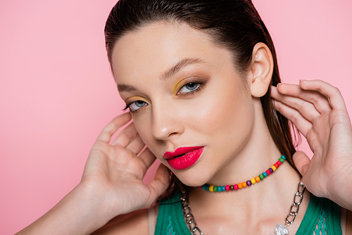 young brunette model with bright makeup posing while looking at camera isolated on pink