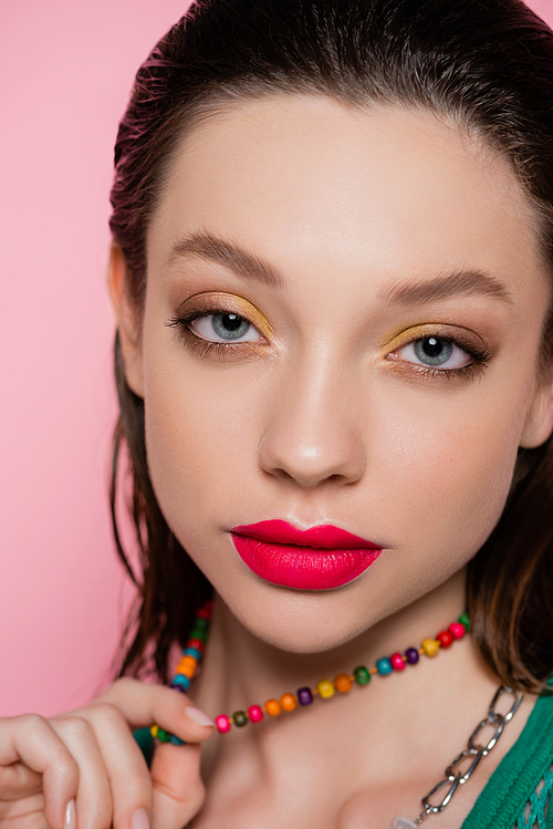 close up view of young model with bright makeup posing while pulling beads necklace isolated on pink
