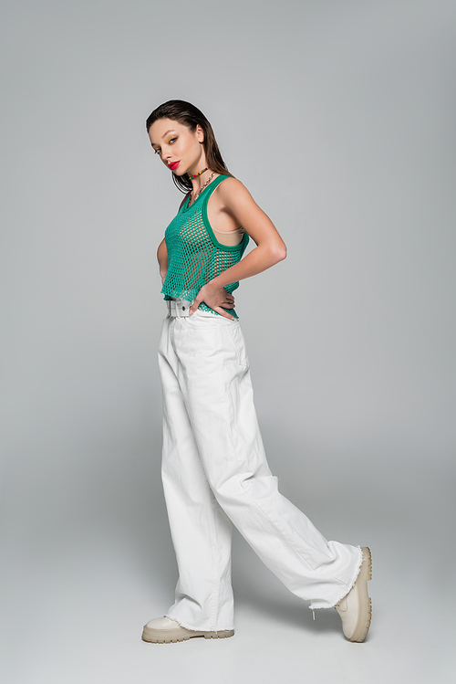 full length of young woman in stylish outfit posing with hand on hip on grey