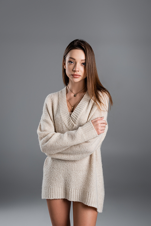 young and pretty woman in sweater and golden necklaces posing with crossed arms on grey background