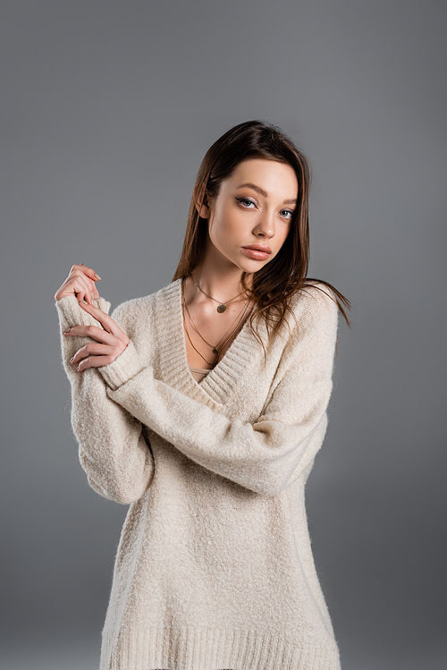 young woman in cozy sweater and golden necklaces looking at camera isolated on grey