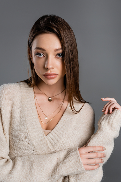 pretty young woman in golden necklaces and soft sweater looking at camera isolated on grey