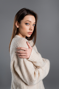 brunette woman in soft sweater hugging herself and looking at camera isolated on grey