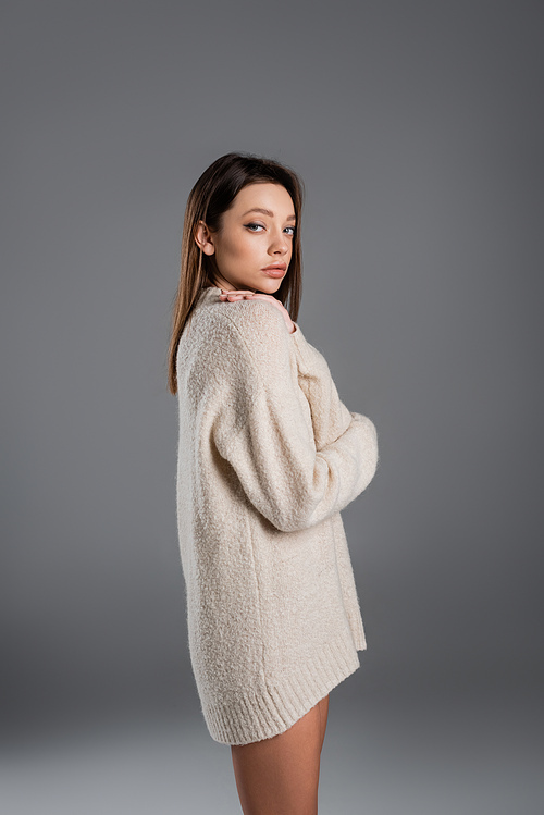 young woman in long and warm sweater looking at camera on grey background