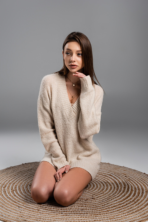 pretty woman in sweater sitting on rug with naked legs and looking away on grey background
