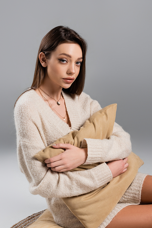 young woman in sweater and golden necklaces hugging pillow on grey background