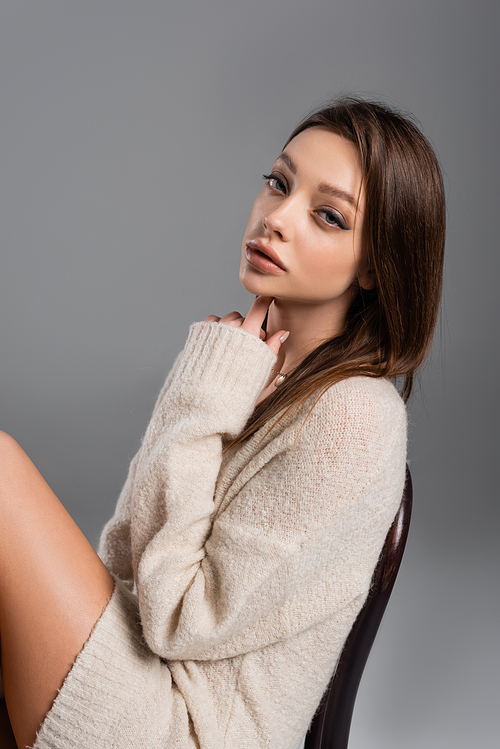sensual woman in cozy sweater touching neck and looking at camera isolated on grey