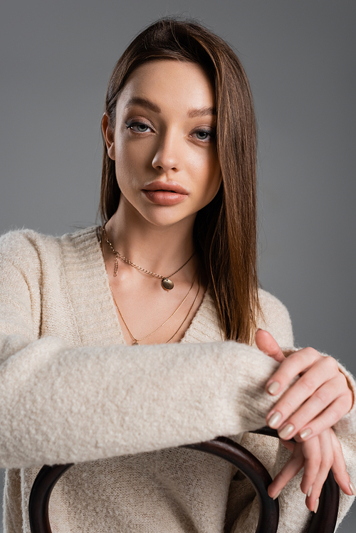 young woman in warm sweater and golden necklaces looking at camera isolated on grey