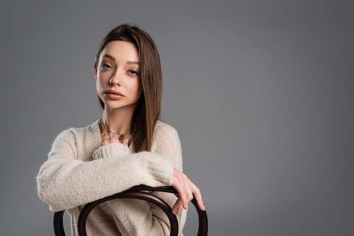 brunette woman in warm sweater looking at camera while posing on chair isolated on grey