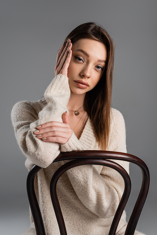 brunette woman in soft sweater sitting on chair and touching face isolated on grey