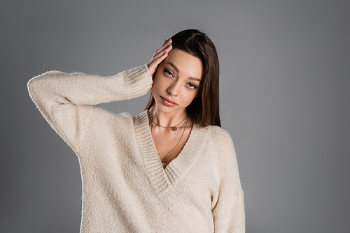 young brunette woman in soft sweater touching head and looking at camera isolated on grey