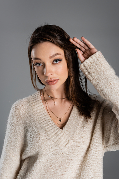 young woman in sweater and golden necklaces touching hair isolated on grey