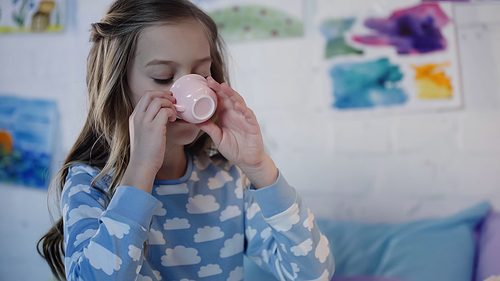 Preteen girl in pajama drinking tea in blurred bedroom