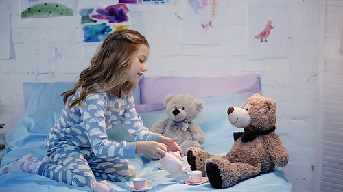 Side view of smiling preteen kid pouring tea near soft toys on bed