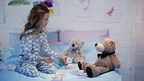 Side view of preteen child in pajama playing near tea cups and soft toys on bed