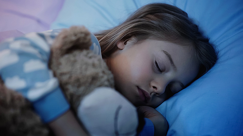 Preteen girl sleeping near blurred soft toy on bed at night