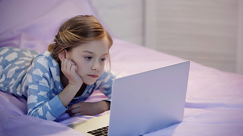Preteen girl in pajama looking at laptop on bed in evening