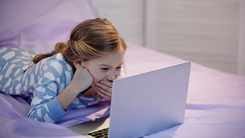 Cheerful preteen kid in pajama looking at laptop while lying on bed