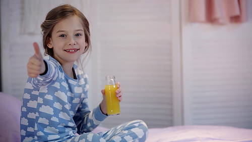 Smiling kid in pajama showing thumb up and holding orange juice on bed