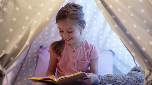 Happy preteen kid reading book in wigwam at home