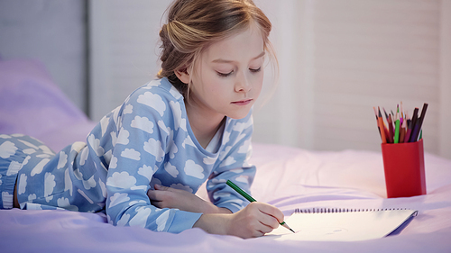 Preteen child in pajama drawing with color pencil on sketchbook on bed