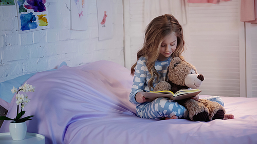 Smiling kid in pajama reading fairytale near soft toy on bed in evening