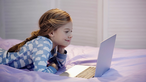 Smiling girl in pajama looking at laptop in bedroom in evening