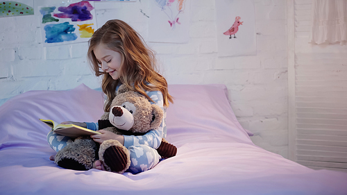 Happy preteen kid reading book and hugging soft toy on bed in bedroom