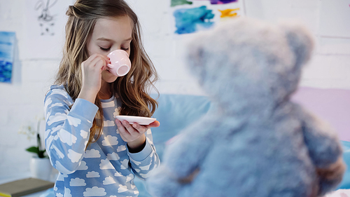 Child in pajama drinking tea near blurred teddy bear in bedroom