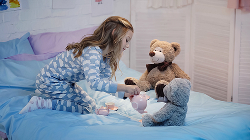 Side view of preteen child pouring tea while playing near teddy bears on bed
