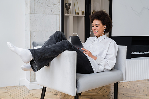 happy african american woman in earphones holding digital tablet while watching movie and sitting on grey armchair