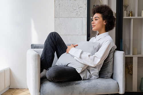 dreamy and curly african american woman in white shirt with collar sitting with pillow on grey armchair