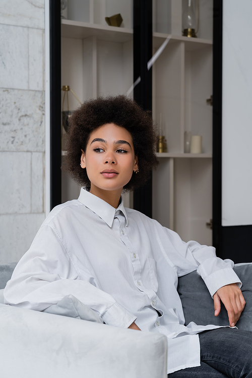 dreamy and curly african american woman in white shirt with collar sitting on grey velvet armchair