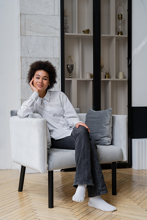 smiling and curly african american woman in white shirt with collar sitting on grey modern armchair