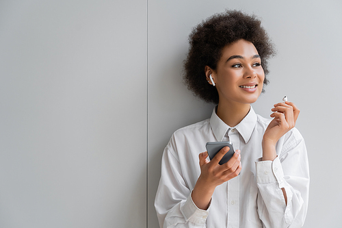 cheerful african american woman listening music in wireless earphone and holding smartphone near grey wall