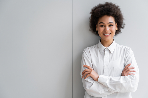 happy african american woman in white shirt with collar standing with crossed arms near grey wall