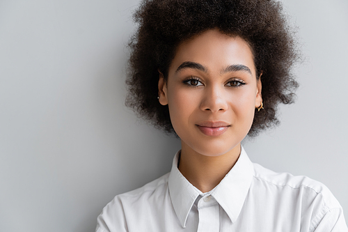 portrait of curly african american woman in white shirt with collar standing near grey wall