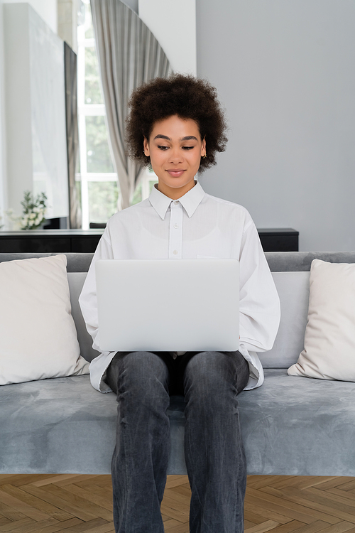 smiling african american woman using laptop while sitting on grey velvet sofa